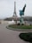 A couple in wedding attire poses in front of the Eiffel Tower. The bride is wearing a white gown with a long train. A photographer is capturing the moment, and the scene includes a statue of a winged horse nearby. Trees and a cloudy sky form the backdrop.