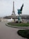 A couple in wedding attire poses in front of the Eiffel Tower. The bride is wearing a white gown with a long train. A photographer is capturing the moment, and the scene includes a statue of a winged horse nearby. Trees and a cloudy sky form the backdrop.