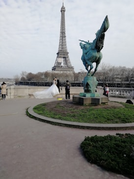 A couple in wedding attire poses in front of the Eiffel Tower. The bride is wearing a white gown with a long train. A photographer is capturing the moment, and the scene includes a statue of a winged horse nearby. Trees and a cloudy sky form the backdrop.