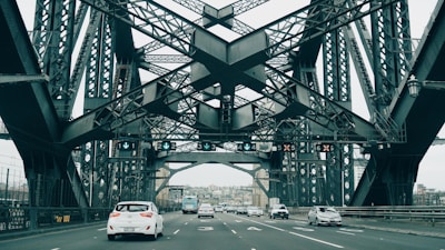 A large steel bridge with intricate latticework spans across a road. Multiple vehicles, including cars and a bus, are visible driving on the road beneath the structure. Overhead signage displays lane directions, and the surrounding environment suggests an urban setting with buildings in the background.
