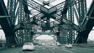 A large steel bridge with intricate latticework spans across a road. Multiple vehicles, including cars and a bus, are visible driving on the road beneath the structure. Overhead signage displays lane directions, and the surrounding environment suggests an urban setting with buildings in the background.