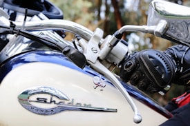 Close-up view of a motorcycle's handlebar with a gloved hand gripping the clutch lever. The motorcycle's logo and model name, Honda Shadow, are prominently displayed on the fuel tank. The background is blurred, highlighting the bike's chrome and detailing.