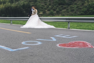A bride in a white wedding dress and a groom in a suit stand together near a metal guardrail. The word 'LOVE' is written in blue on the road with a red heart next to it. The background features greenery and a blurred hillside.
