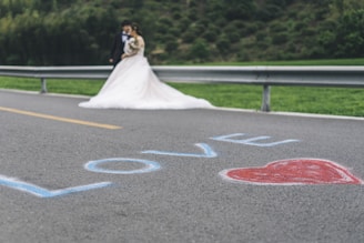 A bride in a white wedding dress and a groom in a suit stand together near a metal guardrail. The word 'LOVE' is written in blue on the road with a red heart next to it. The background features greenery and a blurred hillside.