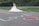 A bride in a white wedding dress and a groom in a suit stand together near a metal guardrail. The word 'LOVE' is written in blue on the road with a red heart next to it. The background features greenery and a blurred hillside.