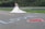 A bride in a white wedding dress and a groom in a suit stand together near a metal guardrail. The word 'LOVE' is written in blue on the road with a red heart next to it. The background features greenery and a blurred hillside.