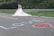 A bride in a white wedding dress and a groom in a suit stand together near a metal guardrail. The word 'LOVE' is written in blue on the road with a red heart next to it. The background features greenery and a blurred hillside.