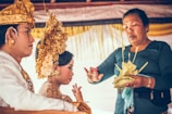 A culturally dressed couple is partaking in a traditional ceremony. The woman is adorned with an elaborate golden headpiece and floral decorations, while the man wears a ceremonial garment. A third person, possibly a celebrant or officiant, is holding a decorative palm leaf arrangement and appears to be conducting a ritual or blessing.