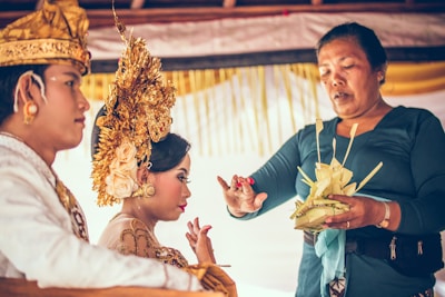 A culturally dressed couple is partaking in a traditional ceremony. The woman is adorned with an elaborate golden headpiece and floral decorations, while the man wears a ceremonial garment. A third person, possibly a celebrant or officiant, is holding a decorative palm leaf arrangement and appears to be conducting a ritual or blessing.