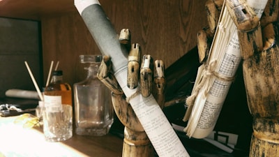 A wooden mannequin hand holds a rolled-up piece of paper with text on it, indicating a theme of writing or literature. Another wooden hand grips a bundle of papers tied with string. In the background, there is a glass bottle and a glass cup on a wooden surface, and a shelf with miscellaneous items.