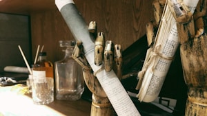 A wooden mannequin hand holds a rolled-up piece of paper with text on it, indicating a theme of writing or literature. Another wooden hand grips a bundle of papers tied with string. In the background, there is a glass bottle and a glass cup on a wooden surface, and a shelf with miscellaneous items.