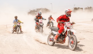 Five motorcyclists are racing on a dusty track, each wearing colorful protective gear. The riders are captured in motion, with a cloud of dust trailing behind them. They are focused and maneuvering around a curve in the track.