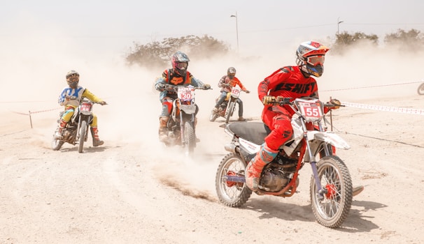Five motorcyclists are racing on a dusty track, each wearing colorful protective gear. The riders are captured in motion, with a cloud of dust trailing behind them. They are focused and maneuvering around a curve in the track.