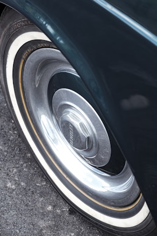 A close-up view of a vintage car wheel featuring a chrome hubcap with the brand name visible. The tire has a classic whitewall design, and the surface beneath the car is asphalt.