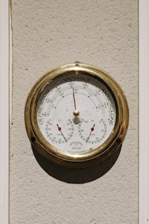 A round antique-style barometer with a brass frame mounted on a textured wall. The instrument displays pressure readings ranging from 'Depression' to 'Anticyclone' with a needle pointing to the current pressure. Below the main dial, two smaller gauges indicate temperature and humidity with red needles.