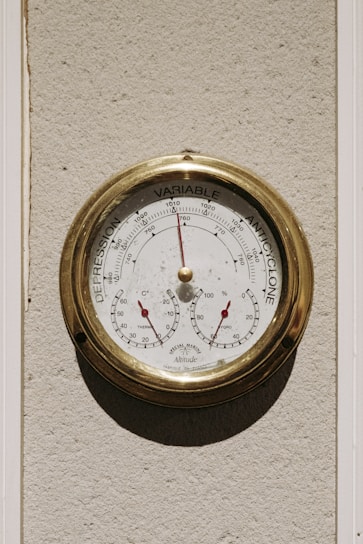 A round antique-style barometer with a brass frame mounted on a textured wall. The instrument displays pressure readings ranging from 'Depression' to 'Anticyclone' with a needle pointing to the current pressure. Below the main dial, two smaller gauges indicate temperature and humidity with red needles.