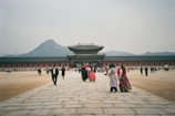 A traditional palace with a wide courtyard and people walking towards the main building. Some individuals are wearing traditional, colorful clothing, adding a cultural element to the scene. The background features a mountain range under a grayish sky.