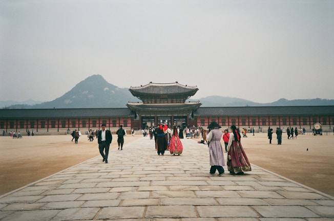 A traditional palace with a wide courtyard and people walking towards the main building. Some individuals are wearing traditional, colorful clothing, adding a cultural element to the scene. The background features a mountain range under a grayish sky.