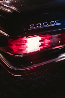 The image focuses on the rear of a vintage Mercedes-Benz 230 CE with distinct tail lights illuminated under low light conditions. The car's dark exterior exudes a classic and luxury vibe, while the model designation is prominently displayed.