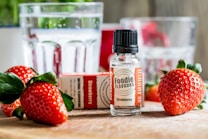 A bottle labeled 'Foodie Flavours Strawberry' stands prominently on a wooden surface surrounded by fresh strawberries and their green leaves. In the background, there are clear glasses containing water, with some out-of-focus reflections, adding a bright and fresh atmosphere.
