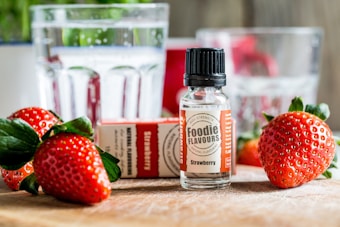 A bottle labeled 'Foodie Flavours Strawberry' stands prominently on a wooden surface surrounded by fresh strawberries and their green leaves. In the background, there are clear glasses containing water, with some out-of-focus reflections, adding a bright and fresh atmosphere.