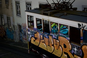 A vintage tram covered in colorful graffiti travels along tracks beside a graffiti-laden building wall. The scene captures urban art and street culture, with vibrant spray-painted designs and tags covering most visible surfaces.