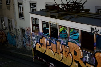 A vintage tram covered in colorful graffiti travels along tracks beside a graffiti-laden building wall. The scene captures urban art and street culture, with vibrant spray-painted designs and tags covering most visible surfaces.