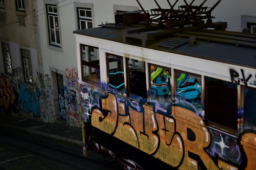A vintage tram covered in colorful graffiti travels along tracks beside a graffiti-laden building wall. The scene captures urban art and street culture, with vibrant spray-painted designs and tags covering most visible surfaces.