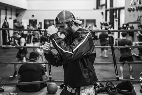 A man in a boxing stance is in focus, wearing a cap and athletic clothing inside a gym. The background is blurred, showing several people engaged in various activities, possibly training or exercising. Boxing equipment such as ropes and punching bags can be seen around.