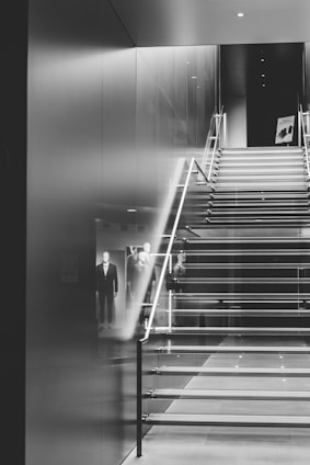 A sleek, modern staircase with glass sides and metal railings, leading upwards in a stylish, minimalistic interior. Reflections and mannequins in suits are visible, indicating a store or showroom setting.