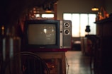 A vintage television set sits on a wooden stand in a dimly lit room. The TV has two dials and a small brand logo on the bottom front. Soft ambient light comes through a window with grid patterns in the background, while various indistinct objects decorate the area, giving a cozy, nostalgic atmosphere.