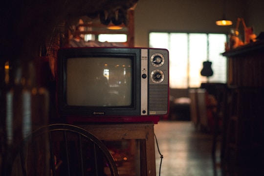 A vintage television set sits on a wooden stand in a dimly lit room. The TV has two dials and a small brand logo on the bottom front. Soft ambient light comes through a window with grid patterns in the background, while various indistinct objects decorate the area, giving a cozy, nostalgic atmosphere.