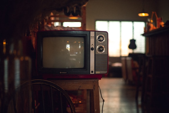 A vintage television set sits on a wooden stand in a dimly lit room. The TV has two dials and a small brand logo on the bottom front. Soft ambient light comes through a window with grid patterns in the background, while various indistinct objects decorate the area, giving a cozy, nostalgic atmosphere.