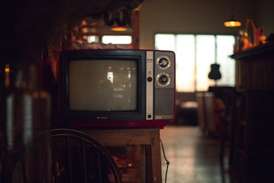 A vintage television set sits on a wooden stand in a dimly lit room. The TV has two dials and a small brand logo on the bottom front. Soft ambient light comes through a window with grid patterns in the background, while various indistinct objects decorate the area, giving a cozy, nostalgic atmosphere.