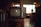 A vintage television set sits on a wooden stand in a dimly lit room. The TV has two dials and a small brand logo on the bottom front. Soft ambient light comes through a window with grid patterns in the background, while various indistinct objects decorate the area, giving a cozy, nostalgic atmosphere.
