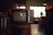 A vintage television set sits on a wooden stand in a dimly lit room. The TV has two dials and a small brand logo on the bottom front. Soft ambient light comes through a window with grid patterns in the background, while various indistinct objects decorate the area, giving a cozy, nostalgic atmosphere.