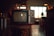 A vintage television set sits on a wooden stand in a dimly lit room. The TV has two dials and a small brand logo on the bottom front. Soft ambient light comes through a window with grid patterns in the background, while various indistinct objects decorate the area, giving a cozy, nostalgic atmosphere.