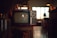 A vintage television set sits on a wooden stand in a dimly lit room. The TV has two dials and a small brand logo on the bottom front. Soft ambient light comes through a window with grid patterns in the background, while various indistinct objects decorate the area, giving a cozy, nostalgic atmosphere.