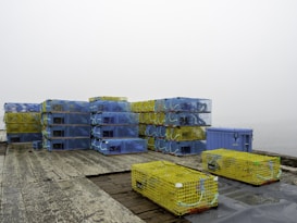 Several stacks of colorful lobster traps are arranged on a weathered wooden dock. The traps are predominantly blue and yellow and are tightly packed together, some with ropes attached. The background features a foggy and indistinct body of water.