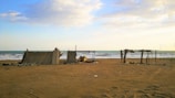 Several makeshift shelters constructed from wood and reeds stand on a sandy beach. The ocean is visible in the background with gentle waves lapping at the shore under a partly cloudy sky. The setting appears calm and somewhat isolated.