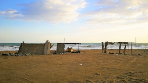 Several makeshift shelters constructed from wood and reeds stand on a sandy beach. The ocean is visible in the background with gentle waves lapping at the shore under a partly cloudy sky. The setting appears calm and somewhat isolated.