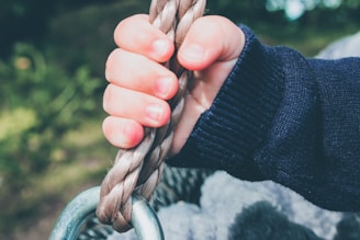 A child's hand grasping a thick rope with a metal ring attached at the bottom, set against a blurred green background. The child is wearing a dark blue sweater.