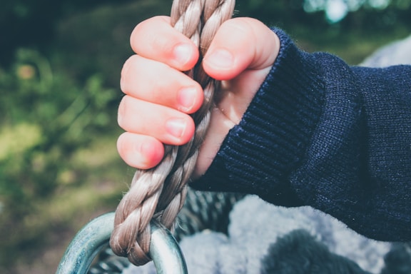 A child's hand grasping a thick rope with a metal ring attached at the bottom, set against a blurred green background. The child is wearing a dark blue sweater.
