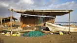 A small, handmade shelter built with wooden poles and thatched roofing stands on a sandy beach. Various fishing gear including nets, buoys, and ropes are scattered around. A white fishing boat is positioned next to the shelter, with the ocean visible in the background. The sky is partially cloudy, casting a mix of light and shadows across the scene.