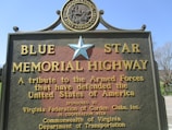 A large, detailed sign commemorating the Blue Star Memorial Highway, paying tribute to the Armed Forces that have defended the United States of America. The sign includes a prominent blue star at the top, with text describing the highway as a tribute. It is sponsored by the Virginia Federation of Garden Clubs, Inc., in cooperation with the Commonwealth of Virginia Department of Transportation.
