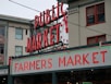 A public market sign and farmers market sign in bright neon lights are displayed against the backdrop of an old building. The red letters are prominent with a large wall clock situated between the words. The building has multiple windows with red frames, contributing to an urban market scene.