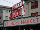A public market sign and farmers market sign in bright neon lights are displayed against the backdrop of an old building. The red letters are prominent with a large wall clock situated between the words. The building has multiple windows with red frames, contributing to an urban market scene.