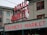 A public market sign and farmers market sign in bright neon lights are displayed against the backdrop of an old building. The red letters are prominent with a large wall clock situated between the words. The building has multiple windows with red frames, contributing to an urban market scene.