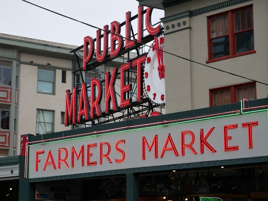 A public market sign and farmers market sign in bright neon lights are displayed against the backdrop of an old building. The red letters are prominent with a large wall clock situated between the words. The building has multiple windows with red frames, contributing to an urban market scene.