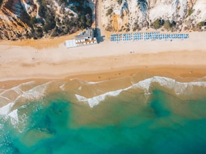 Aerial view of a sandy beach with clear blue water and gentle waves meeting the shore. Sunbeds and umbrellas are arranged neatly in rows, indicating a well-organized beach setup. Cliffs with vegetation rise at one end of the beach, adding a natural backdrop to the scene.